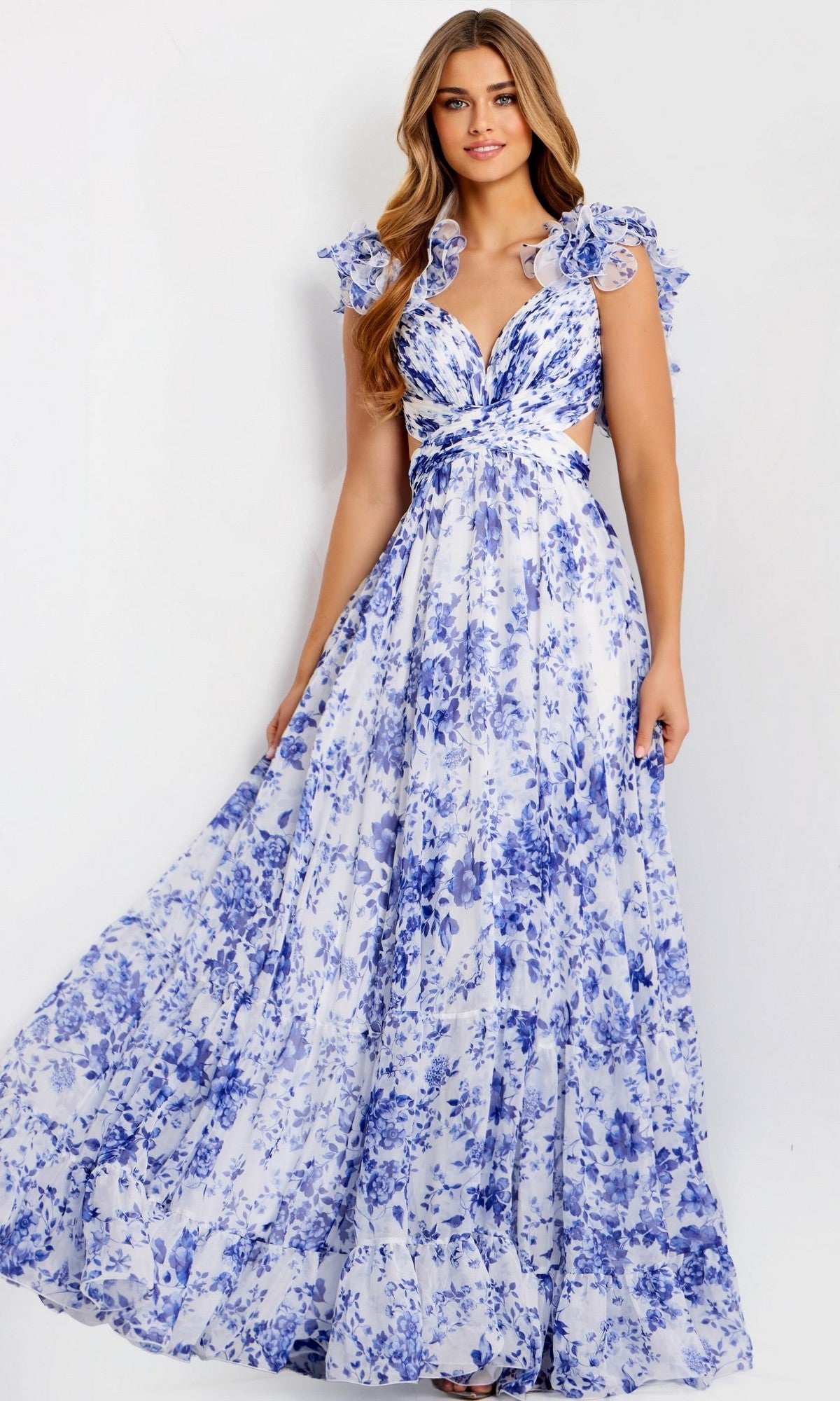 A woman wears the Jovani 49143 Ruffled Long Print Prom Dress—white with blue floral patterns, cap sleeves, ruffles, knotted waist, and a v-neckline—standing against a plain background.